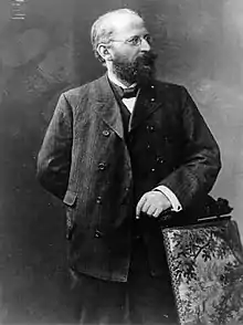 Eduard Bernstein standing next to a chair and looking rightward. He is resting his hand on the chair. (from Socialism)
